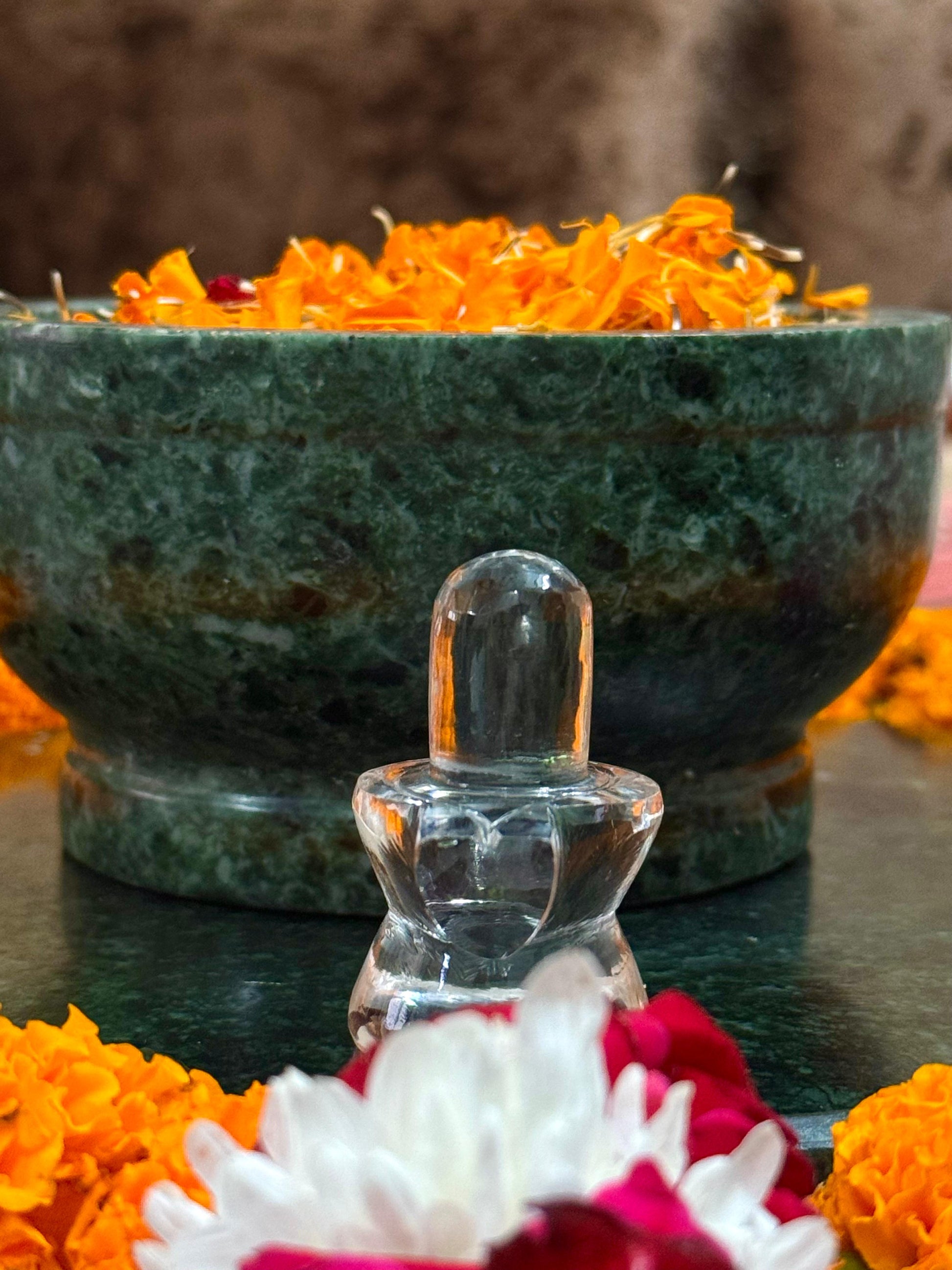 Clear crystal object in front of a large green bowl with marigold flowers on a reflective surface.