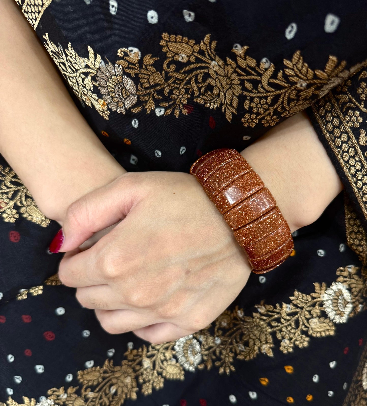 Brown sun stone bracelet on a wrist with a dark fabric background featuring gold embroidery