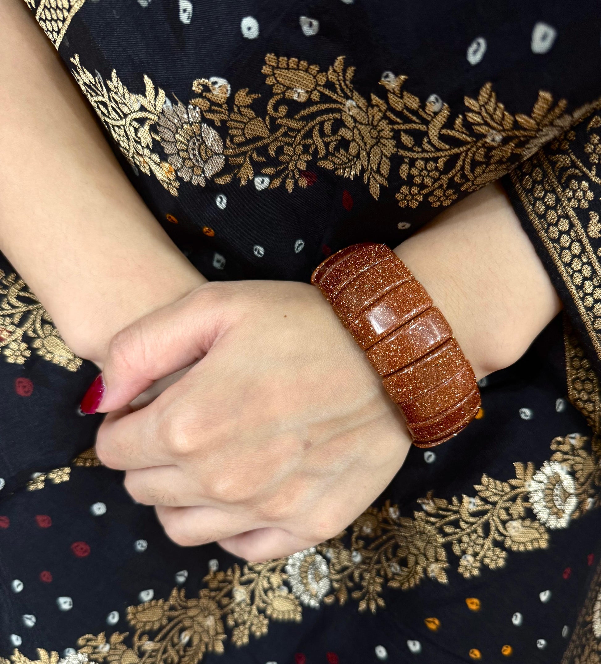 Brown sun stone bracelet on a wrist with a dark fabric background featuring gold embroidery