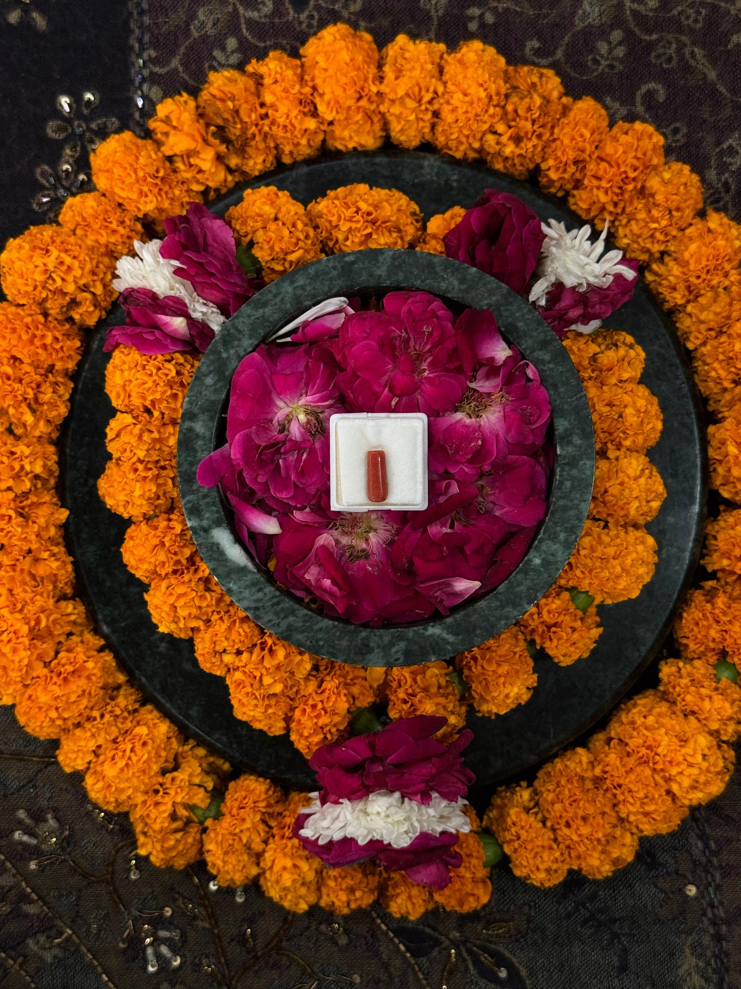 Decorative wreath with marigold flowers, pink and white flowers, and a small red coral stone in the center.
