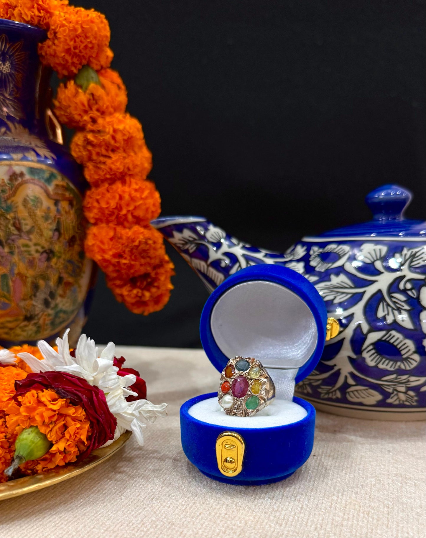 Blue jewelry box with a ring featuring colorful gemstones, surrounded by decorative flowers and a teapot.
